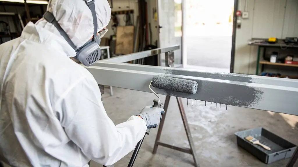 Un professionnel en combinaison de protection applique une couche de primaire antirouille au rouleau sur une rampe en acier décapée.